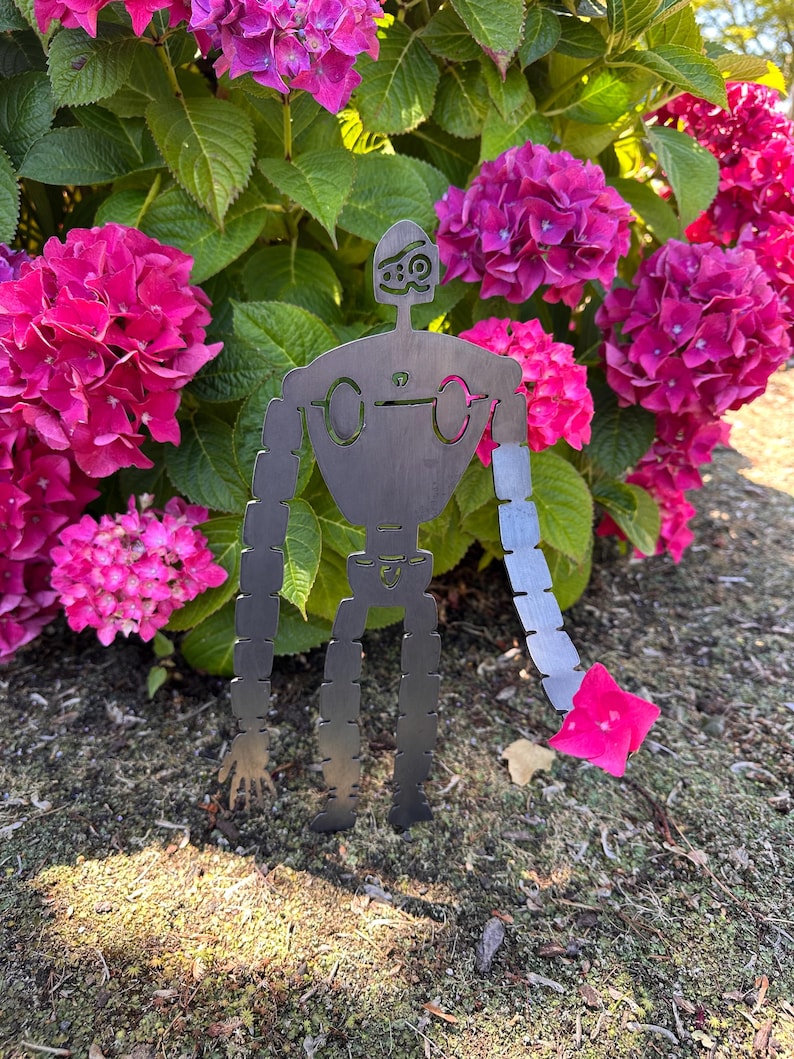 May include: A metal robot garden art piece with a stylized design, standing in front of vibrant pink hydrangea flowers. The robot has a segmented arm holding a pink flower, and is made of dark metal.