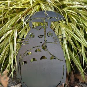 May include: A dark gray metal garden sculpture of a cartoon character holding an umbrella. The sculpture has cut-out details and is placed in front of green and yellow foliage. The sculpture is approximately 18 inches tall.