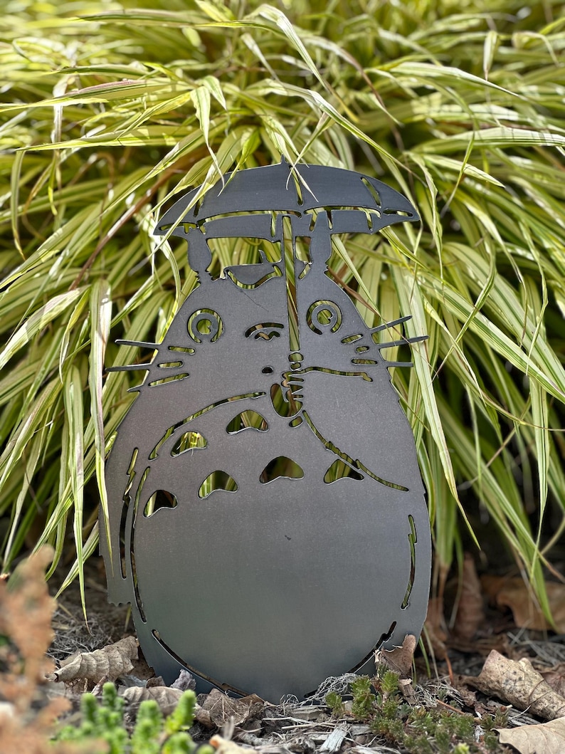 May include: A dark gray metal garden sculpture of a cartoon character holding an umbrella. The sculpture has cut-out details and is placed in front of green and yellow foliage. The sculpture is approximately 18 inches tall.