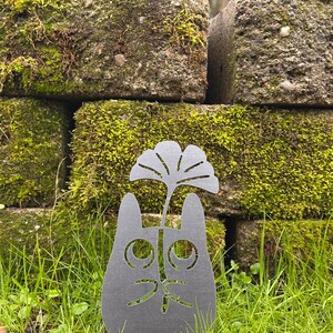 May include: A dark gray metal cat silhouette garden stake with a cut-out face and a stylized leaf design. The stake is placed in green grass in front of moss-covered bricks.