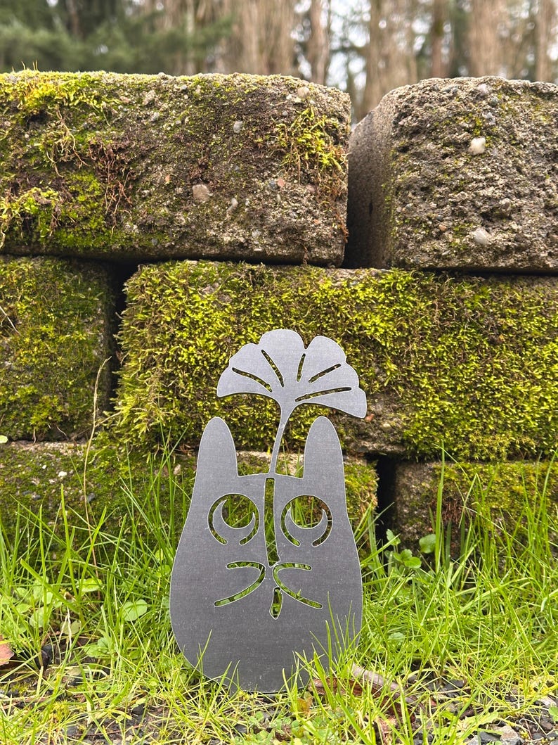 May include: A dark gray metal cat silhouette garden stake with a cut-out face and a stylized leaf design. The stake is placed in green grass in front of moss-covered bricks.
