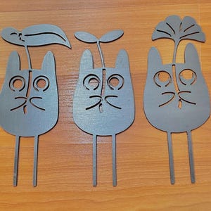 May include: Three gray metal cat-shaped garden stakes with cut-out facial features and plant-like toppers. Each stake has a different leaf or plant design. The stakes are displayed on a wooden surface.