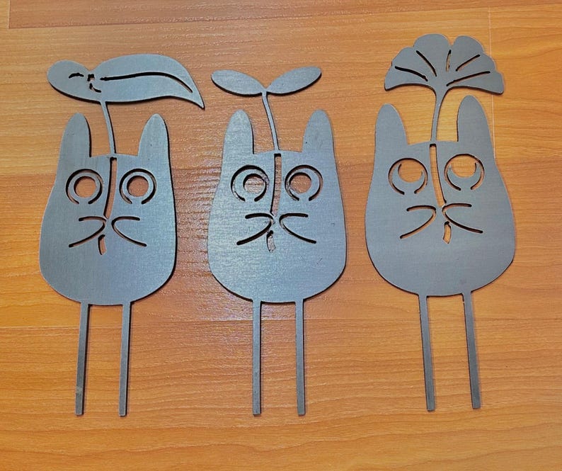 May include: Three gray metal cat-shaped garden stakes with cut-out facial features and plant-like toppers. Each stake has a different leaf or plant design. The stakes are displayed on a wooden surface.