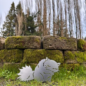 May include: A metal squirrel silhouette, with a fluffy tail and detailed features, sits on green grass in front of a moss-covered brick wall. The squirrel is made of a silver-colored metal and is a decorative garden accent.