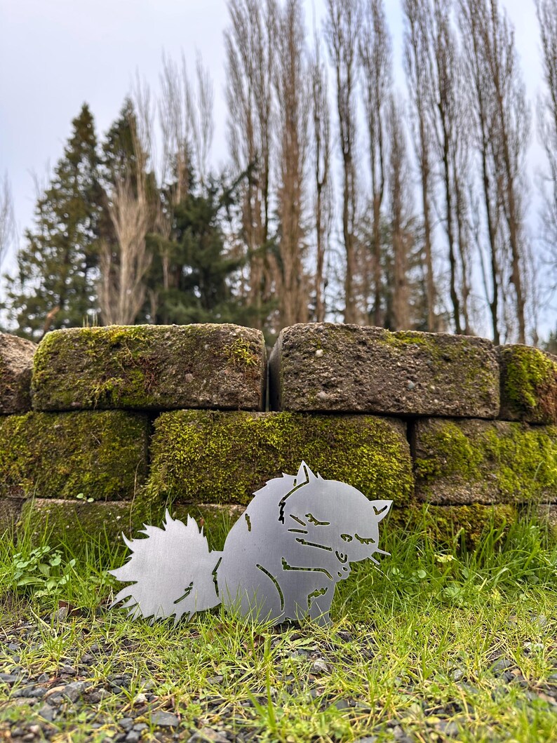 May include: A metal squirrel silhouette, with a fluffy tail and detailed features, sits on green grass in front of a moss-covered brick wall. The squirrel is made of a silver-colored metal and is a decorative garden accent.