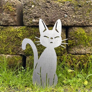May include: A metal cat silhouette garden stake. The cat is sitting with its tail curled and has a simple, minimalist design. The cat is silver and is placed in a grassy area with a mossy brick wall in the background.