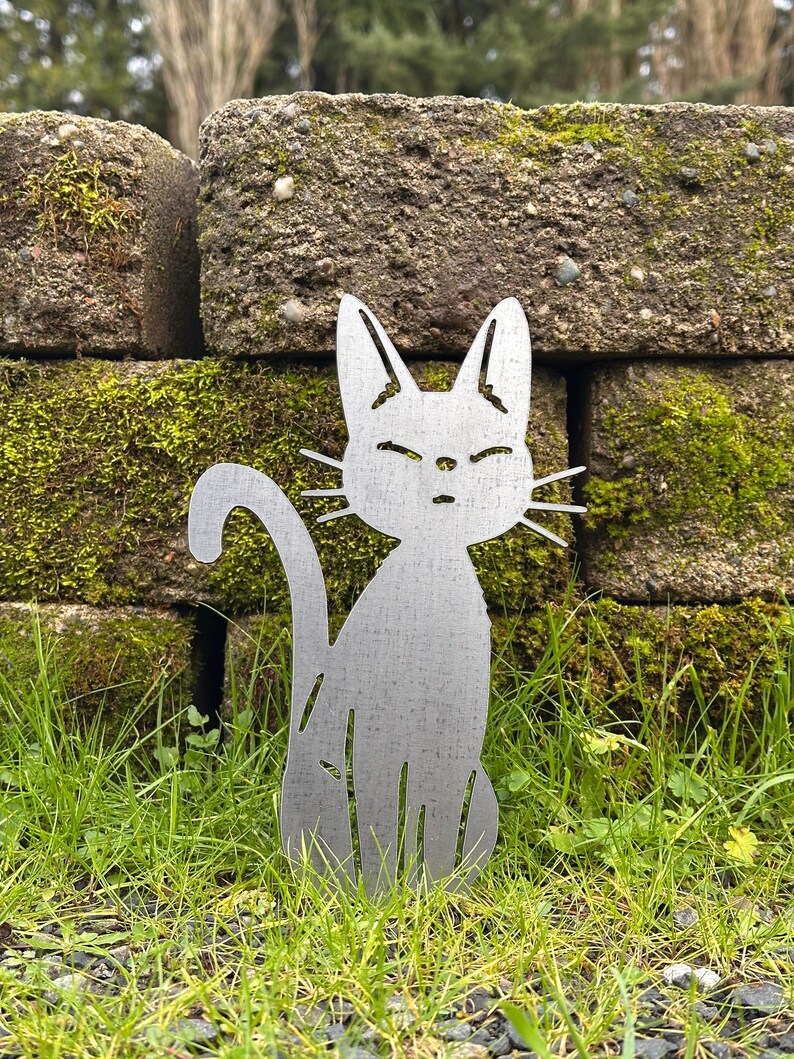 May include: A metal cat silhouette garden stake. The cat is sitting with its tail curled and has a simple, minimalist design. The cat is silver and is placed in a grassy area with a mossy brick wall in the background.