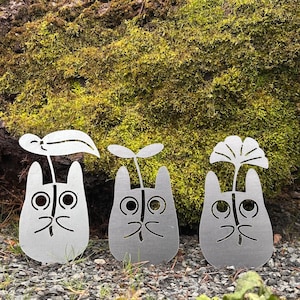 Puede incluir: Tres estacas de jardín de metal plateado con forma de personajes de dibujos animados, cada una con un diseño de planta diferente. Las estacas están colocadas en un lecho de pequeñas rocas, con un fondo cubierto de musgo.
