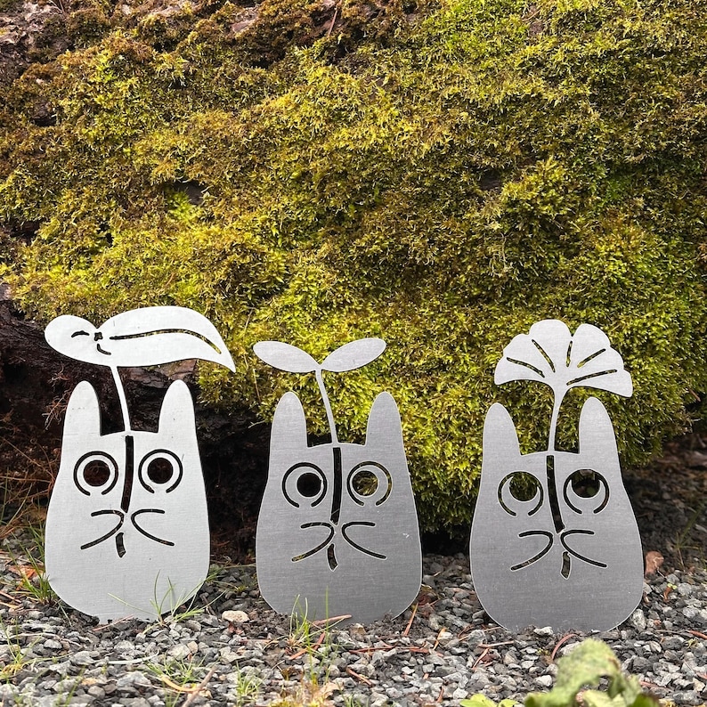 May include: Three silver metal garden stakes shaped like cartoon characters, each with a different plant design sprouting from the top. The stakes are placed in a bed of small rocks with a mossy background.