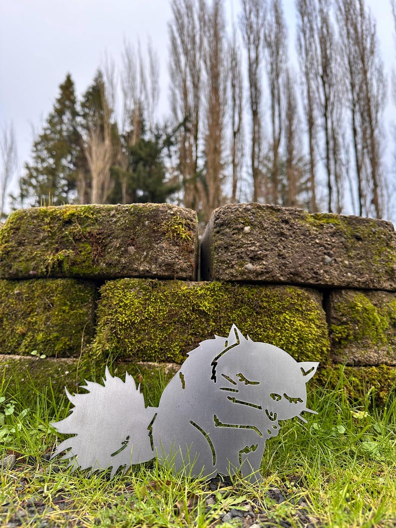 May include: A metal cat silhouette sculpture with a fluffy tail, sitting in green grass. The cat is made of silver metal and is in front of a moss-covered brick wall. The background includes trees and a cloudy sky.