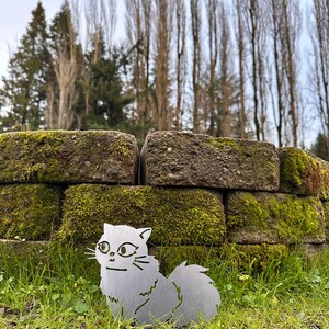 May include: A metal cat silhouette with a fluffy tail sits in green grass in front of a moss-covered brick wall. The cat has detailed facial features and whiskers. Tall trees are in the background.