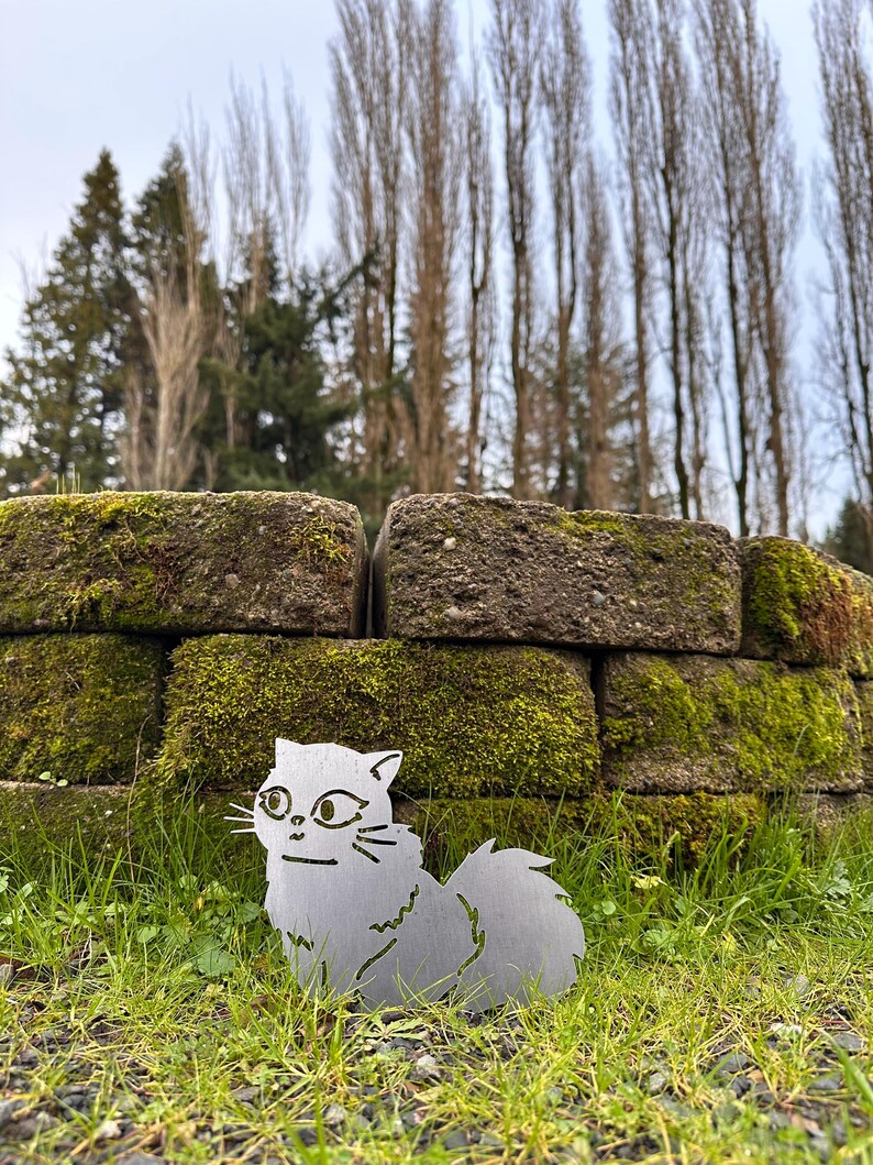 May include: A metal cat silhouette with a fluffy tail sits in green grass in front of a moss-covered brick wall. The cat has detailed facial features and whiskers. Tall trees are in the background.