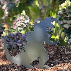 Puede incluir: Silueta metálica de Godzilla, el icónico monstruo de cine, en un jardín. La escultura plateada tiene un contorno detallado, incluyendo las aletas dorsales y la boca abierta. El fondo presenta follaje verde y mantillo marrón.