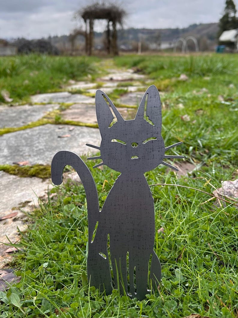 May include: A dark gray metal cat silhouette garden art piece. The cat is sitting upright with its tail curved. The cat is placed in a grassy area with a stone path and a gazebo in the background.