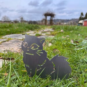 May include: A black metal cat silhouette garden stake. The cat has detailed cutouts for eyes, whiskers, and fur. It is placed in a grassy area with a blurred background of a garden and overcast sky.