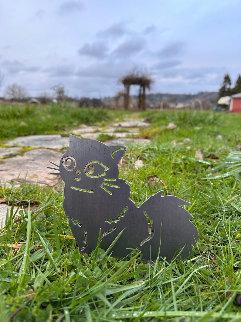 May include: A black metal cat silhouette garden stake. The cat has detailed cutouts for eyes, whiskers, and fur. It is placed in a grassy area with a blurred background of a garden and overcast sky.