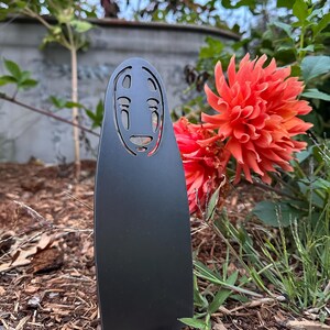 May include: A black metal garden stake featuring a stylized face design. The stake is placed in a garden bed with wood chips, with vibrant orange flowers and green foliage in the background. The design is inspired by a character from an animated film.