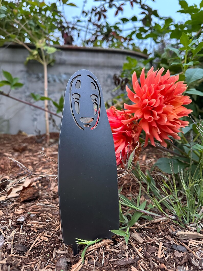 May include: A black metal garden stake featuring a stylized face design. The stake is placed in a garden bed with wood chips, with vibrant orange flowers and green foliage in the background. The design is inspired by a character from an animated film.
