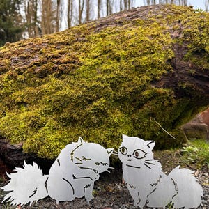 May include: Two metal cat sculptures with detailed features, including fluffy tails and expressive faces, are placed on a bed of gravel. The cats are positioned in front of a moss-covered log, with a blurred forest background.