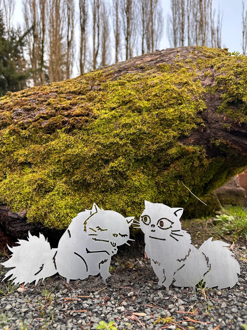 May include: Two metal cat sculptures with detailed features, including fluffy tails and expressive faces, are placed on a bed of gravel. The cats are positioned in front of a moss-covered log, with a blurred forest background.