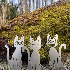May include: Three metal cat yard art sculptures with different facial expressions. The cats are silver and sit on the ground in front of a moss-covered log. The background includes trees and a cloudy sky.