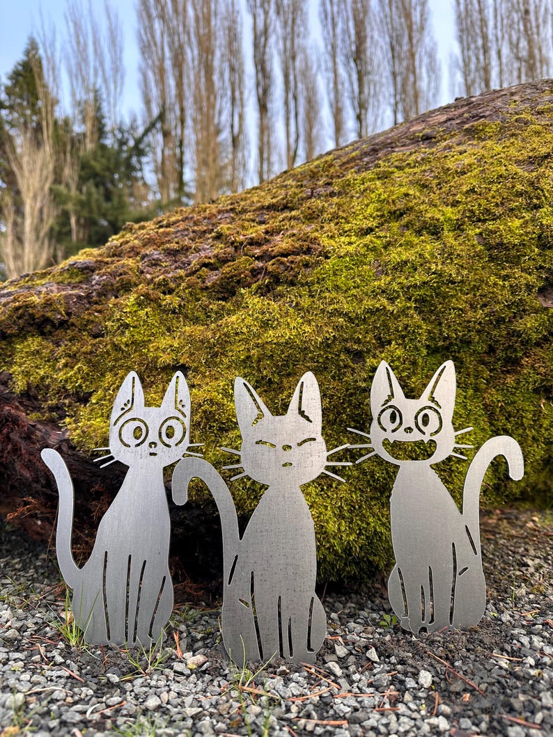 May include: Three metal cat yard art sculptures with different facial expressions. The cats are silver and sit on the ground in front of a moss-covered log. The background includes trees and a cloudy sky.