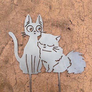 May include: A metal cat garden stake featuring two cats. One cat has large eyes and is sitting upright, while the other cat is curled up next to it. The cats are made of silver metal and have a simple, stylized design.