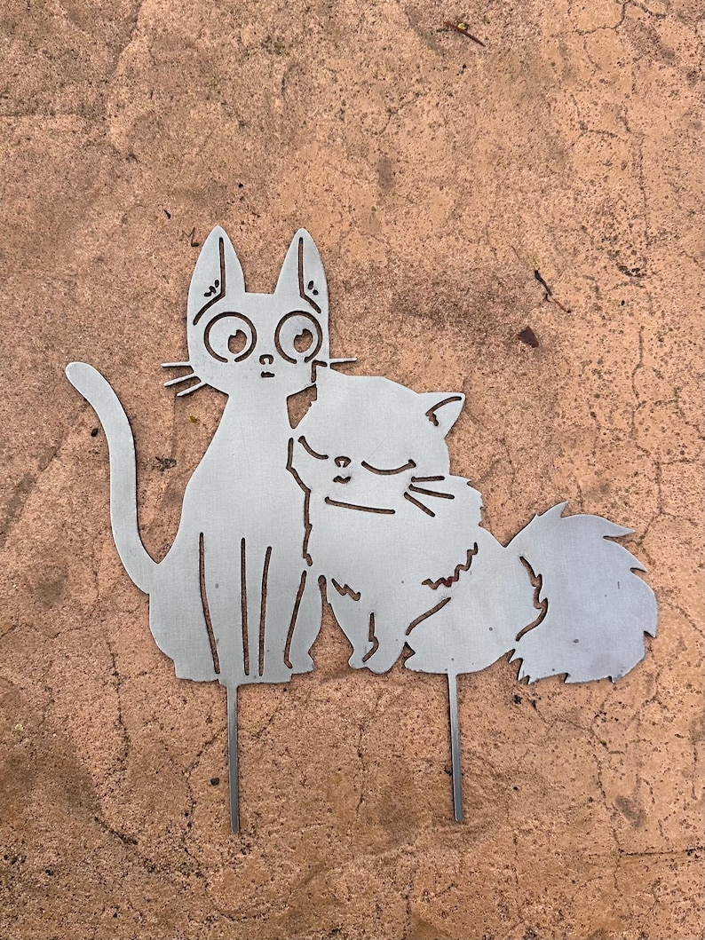 May include: A metal cat garden stake featuring two cats. One cat has large eyes and is sitting upright, while the other cat is curled up next to it. The cats are made of silver metal and have a simple, stylized design.