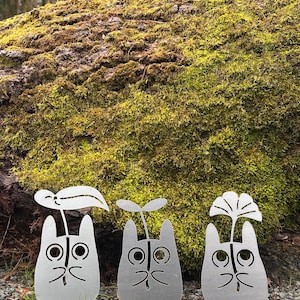 May include: Three metal garden sculptures shaped like cartoon characters with plant-like features. The sculptures are silver and stand on a bed of gravel and grass, with a moss-covered log in the background.