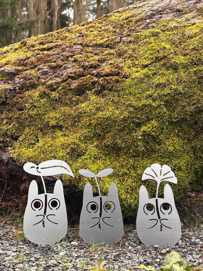 May include: Three metal garden sculptures shaped like cartoon characters with plant-like features. The sculptures are silver and stand on a bed of gravel and grass, with a moss-covered log in the background.