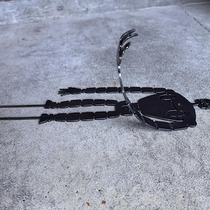 May include: A black metal sculpture of a stylized figure lying down with arms outstretched. The figure has a rounded head and segmented body. The sculpture is placed on a concrete surface.