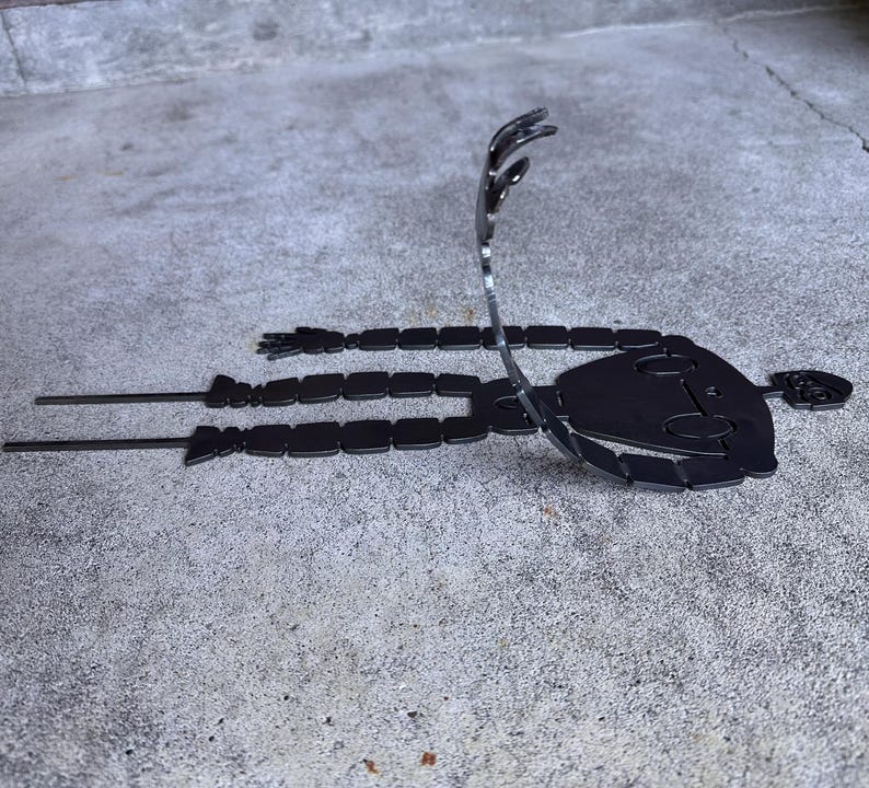 May include: A black metal sculpture of a stylized figure lying down with arms outstretched. The figure has a rounded head and segmented body. The sculpture is placed on a concrete surface.