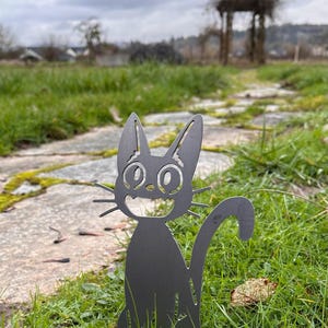 May include: A black metal cat garden stake with a smiling face and long whiskers. The cat is sitting in green grass on a stone path, with a blurred background of a cloudy sky and a wooden arbor.