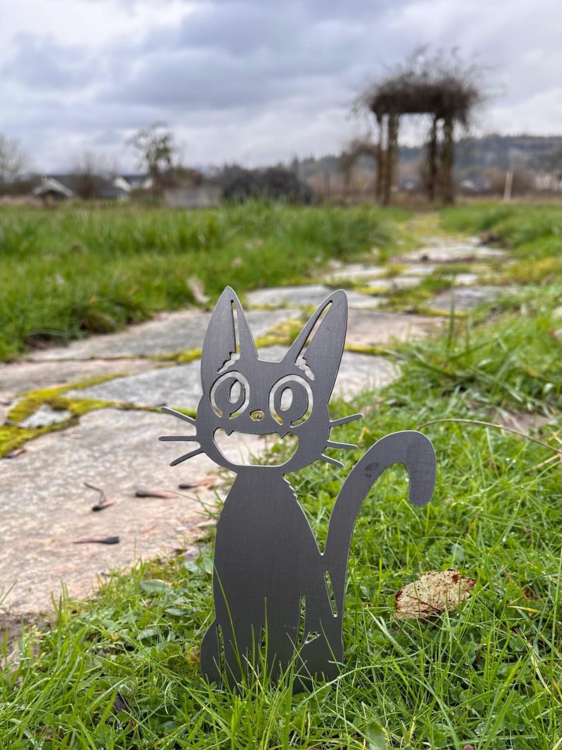May include: A black metal cat garden stake with a smiling face and long whiskers. The cat is sitting in green grass on a stone path, with a blurred background of a cloudy sky and a wooden arbor.