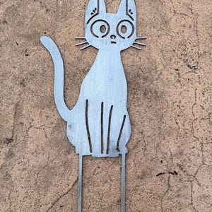 May include: A metal cat garden stake with a whimsical design. The cat is depicted in a seated position with large, round eyes and whiskers. The stake has two prongs for easy placement in the ground. The cat is made of silver metal.