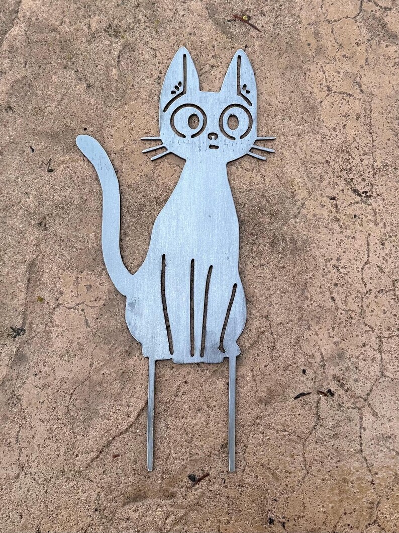 May include: A metal cat garden stake with a whimsical design. The cat is depicted in a seated position with large, round eyes and whiskers. The stake has two prongs for easy placement in the ground. The cat is made of silver metal.