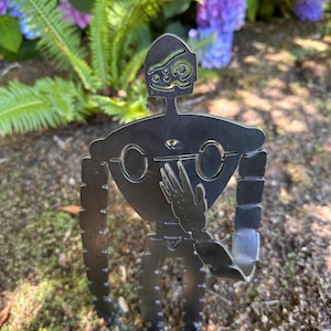 May include: A metal garden art piece depicting a robot figure with a stylized design. The robot has a head with detailed features, a body with circular cutouts, and outstretched arms. The art is placed in a garden setting with purple and blue flowers in the background.