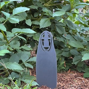 May include: A dark gray metal garden stake featuring a stylized face design. The stake is in a garden setting with green foliage and brown mulch in the background. The design is reminiscent of a character from an animated film.