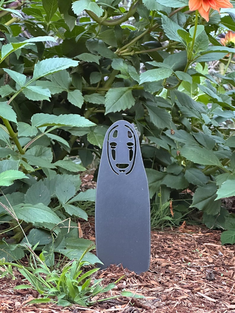 May include: A dark gray metal garden stake featuring a stylized face design. The stake is in a garden setting with green foliage and brown mulch in the background. The design is reminiscent of a character from an animated film.