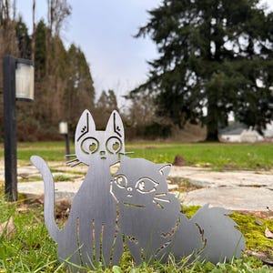 May include: Two metal cat silhouettes, one upright and one cuddling, stand on a grassy surface. The cats are a dark gray color and have detailed cut-out features. The background includes trees and a cloudy sky, suggesting an outdoor garden decoration.
