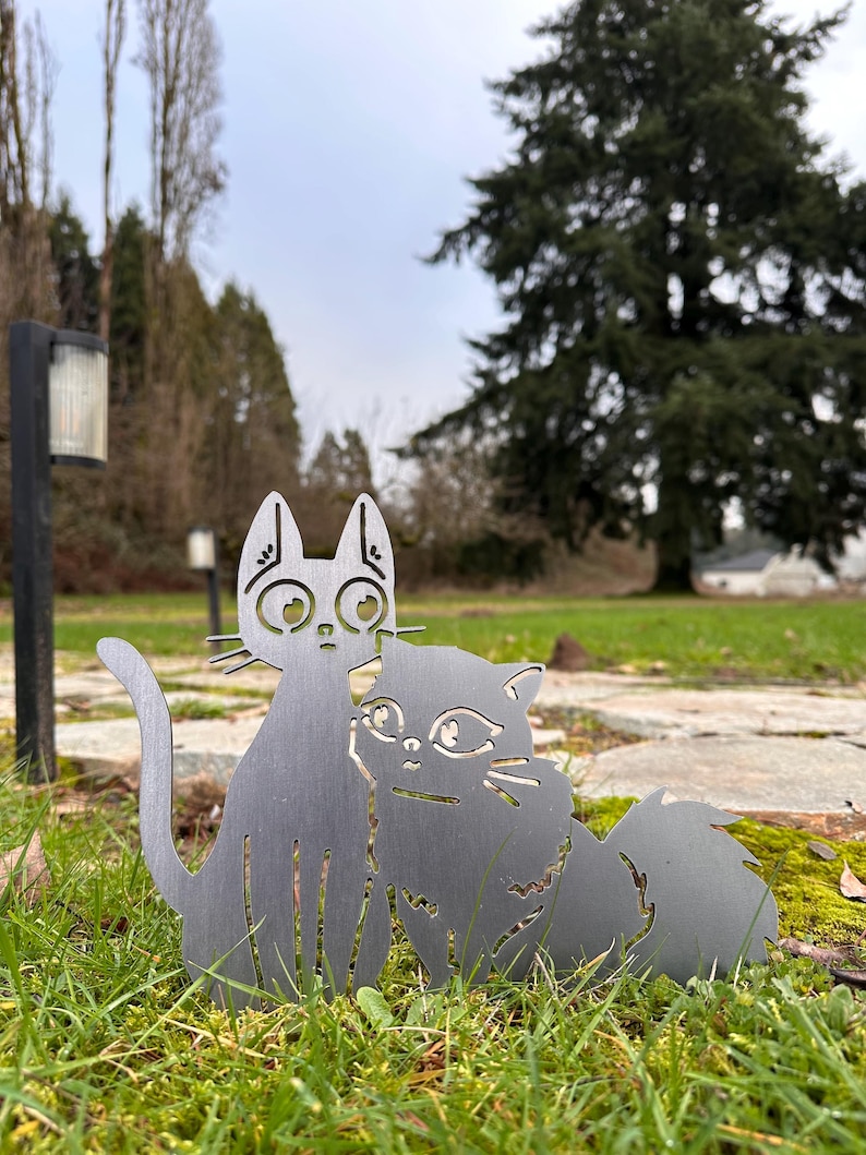 May include: Two metal cat silhouettes, one upright and one cuddling, stand on a grassy surface. The cats are a dark gray color and have detailed cut-out features. The background includes trees and a cloudy sky, suggesting an outdoor garden decoration.