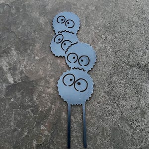 K&ouml;nnte beinhalten: Ein Metall-Gartenstecker mit drei gestapelten, cartoonartigen Figuren. Jede Figur ist rund mit gezackten Kanten, gro&szlig;en Augen und einem kleinen Mund. Die Figuren sind grau mit schwarzen Merkmalen. Zwei schwarze St&auml;be erstrecken sich von der Basis.