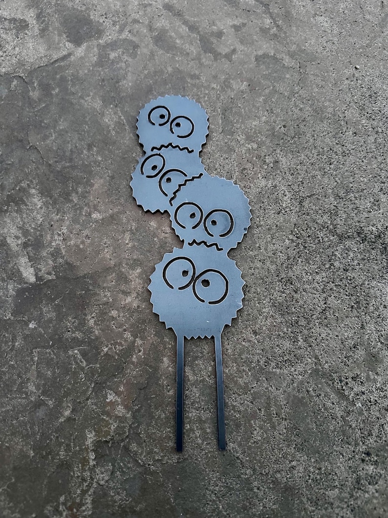 K&ouml;nnte beinhalten: Ein Metall-Gartenstecker mit drei gestapelten, cartoonartigen Figuren. Jede Figur ist rund mit gezackten Kanten, gro&szlig;en Augen und einem kleinen Mund. Die Figuren sind grau mit schwarzen Merkmalen. Zwei schwarze St&auml;be erstrecken sich von der Basis.