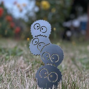 K&ouml;nnte beinhalten: Dunkelgraue Metallgartenkunst mit drei gestapelten, stilisierten Figuren mit gro&szlig;en Augen und gezackten Kanten. Die Figuren stehen in einem grasbewachsenen Bereich mit einem verschwommenen Hintergrund aus Blumen und Bl&auml;ttern.