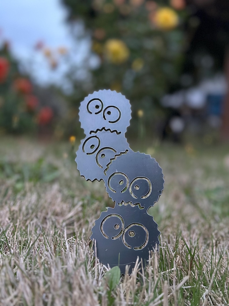 K&ouml;nnte beinhalten: Dunkelgraue Metallgartenkunst mit drei gestapelten, stilisierten Figuren mit gro&szlig;en Augen und gezackten Kanten. Die Figuren stehen in einem grasbewachsenen Bereich mit einem verschwommenen Hintergrund aus Blumen und Bl&auml;ttern.