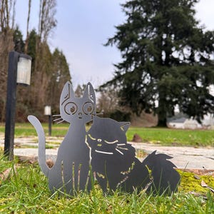 May include: Two metal cat silhouettes, one upright and one seated, are positioned in green grass. The cats are a dark gray color and have cut-out details for eyes, noses, and fur. The background includes trees and a cloudy sky.