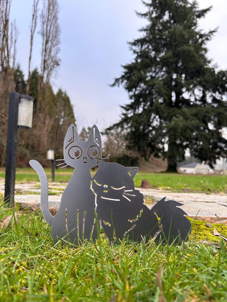May include: Two metal cat silhouettes, one upright and one seated, are positioned in green grass. The cats are a dark gray color and have cut-out details for eyes, noses, and fur. The background includes trees and a cloudy sky.
