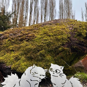 May include: Two metal cat sculptures, one with closed eyes and the other with open eyes, are placed on the ground. The cats are in front of a moss-covered log and tall trees. The sculptures are made of silver metal.