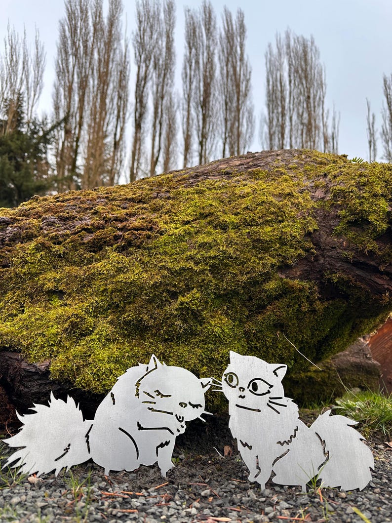 May include: Two metal cat sculptures, one with closed eyes and the other with open eyes, are placed on the ground. The cats are in front of a moss-covered log and tall trees. The sculptures are made of silver metal.