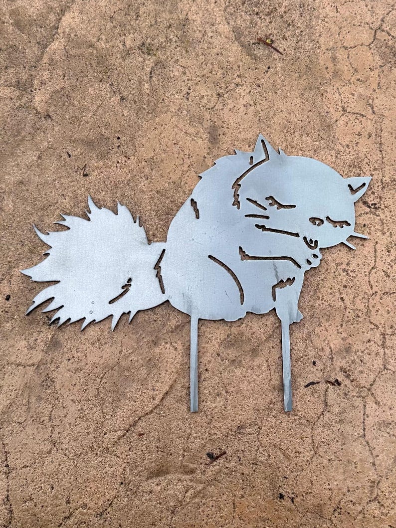 May include: A metal cat silhouette garden stake. The cat is depicted in a sitting position with a fluffy tail and pointed ears. The metal has a silver color and is designed to be placed in the ground.